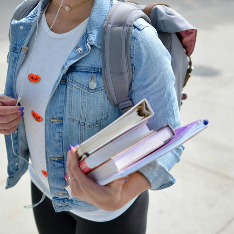 Persona con mochila que sostiene varios libros en una mano. Lleva una camiseta clara.